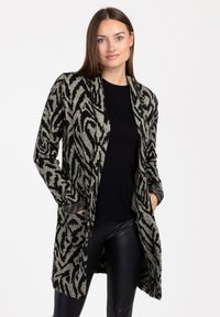 Długi kardigan w kolorze czarnym i beżowym, z abstrakcyjnym wzorem, otwarty przód, obniżone ramiona i kieszenie. Nosi się go na czarną bluzkę i spodnie.