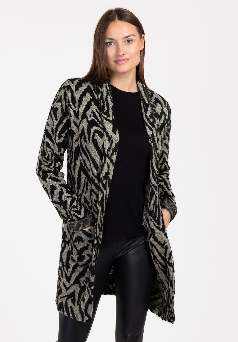 Długi kardigan w kolorze czarnym i beżowym, z abstrakcyjnym wzorem, otwarty przód, obniżone ramiona i kieszenie. Nosi się go na czarną bluzkę i spodnie.