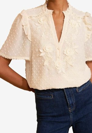 Crèmekleurige blouse met bloemige kanten borduursel, korte pofmouwen en gerimpelde kraag, gedragen met donkerblauwe hoge taille jeans.