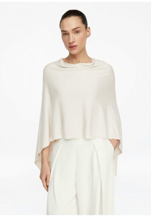 Kobieta z włosami zaczesanymi do tyłu, ubrana w luźne, kremowe ponczo na białych spodniach z wysokim stanem na jednolitym tle.