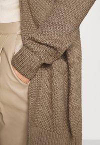 Pletený kardigan světle hnědé barvy s texturovaným vzorem; má volný střih a žebrované manžety, doplněný béžovými koženými kalhotami.