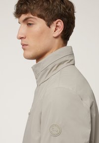 Chaqueta gris claro con cuello alto, detalle de cremallera en el frontal y un parche de logo redondo en la manga izquierda. Fabricada con un material suave y ligero.