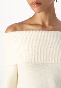 Κρέμα ζακέτα με ανοιχτούς ώμους και πλισέ άνω πάνελ, μαλακή υφή και χαλαρή εφαρμογή, αναδεικνύοντας το λαιμό και την κλείδα.