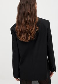 Black blazer featuring a tailored fit, shoulder pads, and a smooth texture. Long sleeves and a center back seam detail.