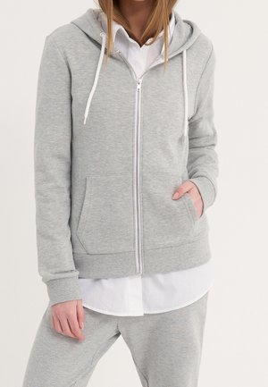Žena oblečená v světle šedé mikině na zip přes bílou košili s knoflíky a odpovídající šedé kalhoty, stojící s jednou rukou v kapse.