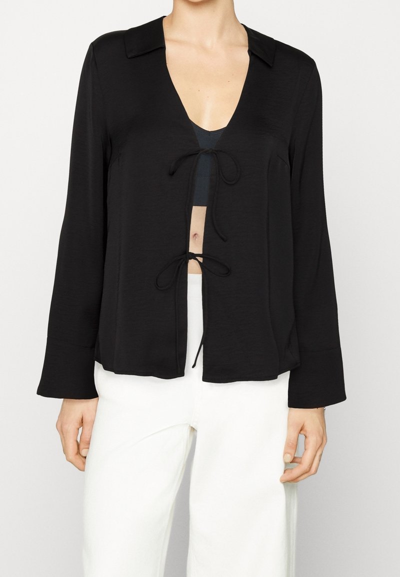 Blouse noire à manches longues avec un décolleté en V et un détail de noeud à l'avant. Fabriquée en tissu léger avec une texture douce. Manches évasées et design ouvert à l'avant.