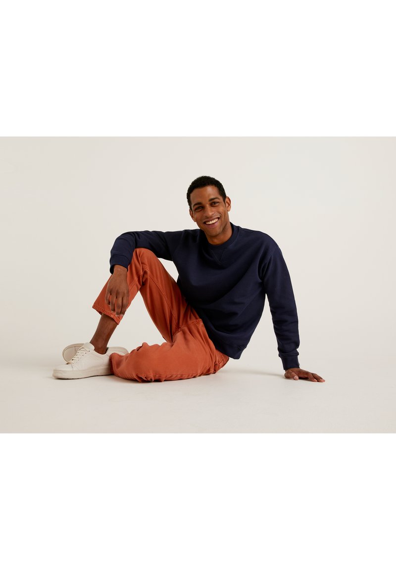Sudadera azul marino, pantalones naranjas y zapatillas blancas. El modelo está sentado en el suelo, sonriendo, con una pose relajada. Fondo simple.