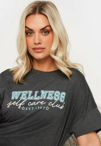 T-shirt grigio a maniche corte con la scritta "WELLNESS self care club" in lettere blu e bianche. Materiale morbido con una vestibilità rilassata, adatto per un abbigliamento informale.