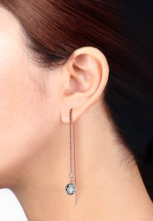 Boucles d'oreilles en or rose avec une longue chaîne qui se termine par un cristal gris façonné, pendent élégamment à côté de l'oreille.