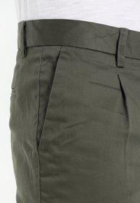 Pantaloni verde oliva realizzati in tessuto liscio, con una parte anteriore piatta, tasche laterali e passanti per la cintura, con una vestibilità su misura e dettagli minimi.