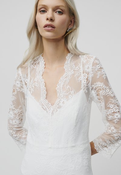 Robe de mariée blanche avec une superposition en dentelle à motifs floraux, manches longues en transparent, et décolleté en V. Corsage texturé avec des détails complexes.