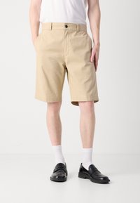 Beige shorts med knappelukking, som har to sidelommer og rett snitt, brukt sammen med sorte loafers og hvite sokker.