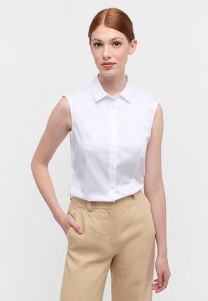 Camicia bianca senza maniche con bottoni davanti e colletto, abbinata a pantaloni beige chiaro, realizzati in un morbido tessuto misto cotone.