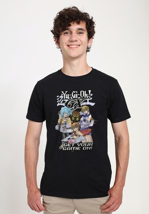 Jonge man die een zwart Yu-Gi-Oh! GX T-shirt draagt met drie animepersonages, een uil en de tekst "Get Your Game On!"