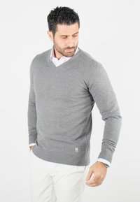 Pull gris en V en tissu doux, avec des manches longues et des poignets côtelés. Accent logo près de l'ourlet, superposé sur une chemise blanche.