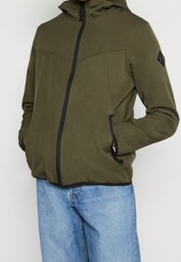 Oliivinvihreä hupullinen takki, jossa etuvetoketju, mustat yksityiskohdat ja sivutaskut; sileä pinta ja istuva malli.