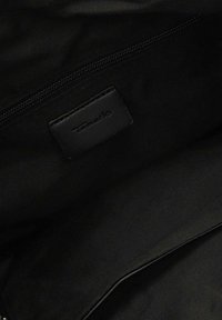 Schwarzer Stoffinnenraum einer Tasche mit einer Reißverschlusstasche und einem aufgenähten rechteckigen Etikett mit eingeprägtem Markennamen.