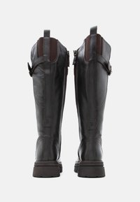 Pitkät mustat nahkakengät ruskeilla yksityiskohdilla, joissa on teksturoitu yläosa, sivuvetoketjut ja kestävä kumipohja. Kuusikulmainen logo yksityiskohta sivuilla.