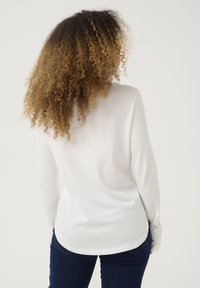 Langärmliges weißes Oberteil aus weichem Stoff, mit abgerundetem Saum und lockerer Passform, kombiniert mit dunkelblauen Jeans.