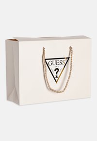 Busta da shopping bianca con manici in corda dorata attorcigliata e un logo triangolare nero con la scritta "GUESS" e "U.S.A." stampata su di essa.
