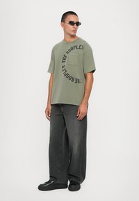 T-shirt de algodão verde oliva com um bolso redondo e texto circular preto. Combinado com jeans de denim pretos de corte largo e sapatos pretos.