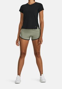 Nainen, jolla on päällään musta lyhythihainen paita, oliivinvihreät urheilushortsit mustilla yksityiskohdilla ja valkoiset juoksukengät, seisoo tasaisella taustalla.