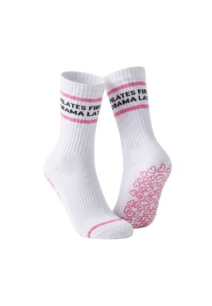 Nogi w białych rajstopach ze skrzyżowanymi stopami, ubrane w białe skarpetki z różowymi paskami i czarnym tekstem, stojące na jednolitym jasnym tle.