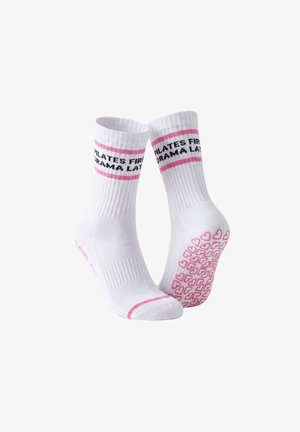 Nogi w białych rajstopach ze skrzyżowanymi stopami, ubrane w białe skarpetki z różowymi paskami i czarnym tekstem, stojące na jednolitym jasnym tle.