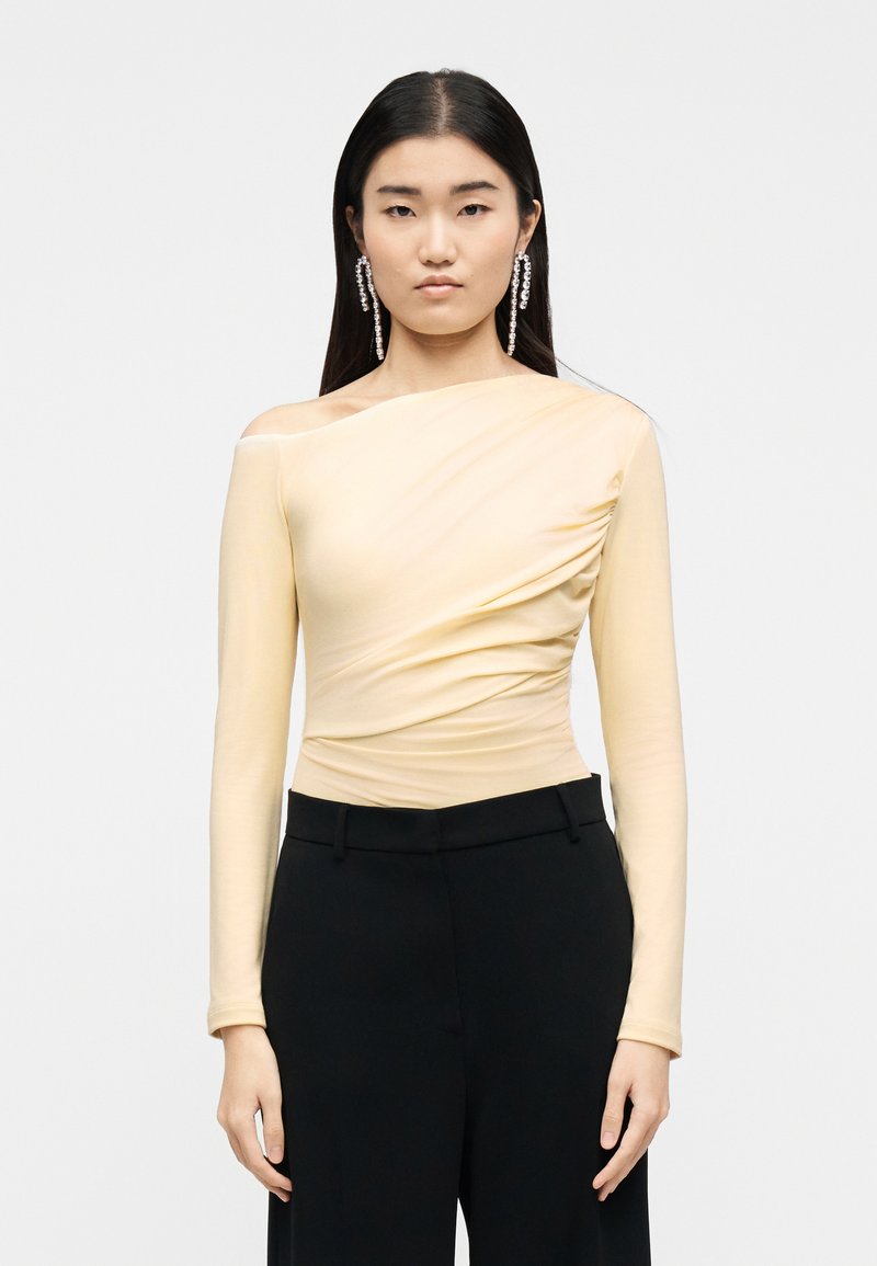 Γυναίκα με μακριά μαύρα μαλλιά, που φοράει ανοιχ τό κίτρινο τοπ με έναν ώμο, μαύρο παντελόνι και μακριά σκουλαρίκια με κρυστάλλους, σε λευκό φόντο.