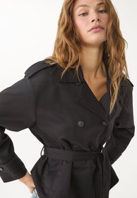 Svart trenchcoat med dubbelknäppt design, axelklaffar och bälte i midjan. Slät överdel med klassisk krage och långa ärmar.