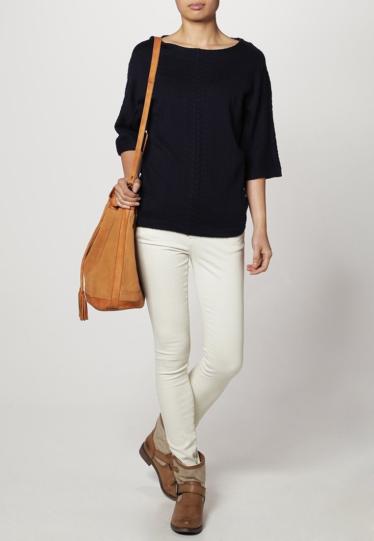 Suéter de punto azul marino, corte holgado, cuello redondo; jeans ajustados crema; botas de tobillo marrones; bolsa de hombro de ante marrón con detalles de flecos.