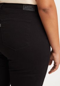 Jean en denim noir avec une taille haute, poches arrière dotées d'une couture courbée, et une patte en cuir texturé à la ceinture arrière.