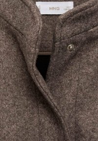 Bruine wollen jas met een zachte, gestructureerde afwerking, voorzien van een kraag, zichtbare drukknoopsluitingen en een eenvoudig, strak ontwerp. Labels aan de binnenkant.