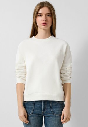 Weißes Baumwoll-Sweatshirt mit rundem Ausschnitt und langenÄrmeln, das ein kleines QS-Logo auf der Brust trägt. Kombiniert mit blauen Jeans.
