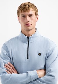 Lichtblauwe fleece pullover met een ritskraag, zwarte rits en een hexagonaal logo op de linkerkant van de borst. Zachte textuur, casual ontwerp.