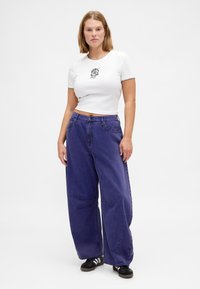 Bela krajšana majica s črnimi poudarki in grafično zasnovo; ohlapne vijoličaste kavbojke z našitki; črni supergi.