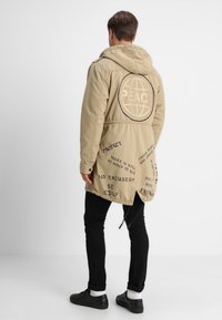 Beżowa kurtka z kapturem z teksturowanej tkaniny, z logo "PEACE" na plecach oraz różnymi nadrukowanymi napisami po bokach. Czarne spodnie i buty.