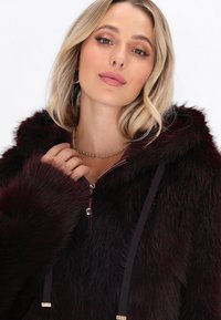 Blondīne ar vieglu grimu uzliek tumši burgundijas krāsas mākslīgā kažoka jaku ar rāvējslēdzēju un tur apkakli netālu no pagrieztās ķēdes kaklarotas.