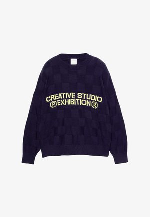 Mornarska pulover z strukturiranim karo vzorcem in okroglo ovratnikom. Na prsih je napis "KREATIVNI STUDIO RAZSTAVA" v rumeni barvi.