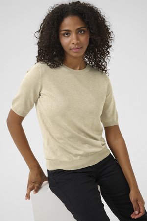Femme aux cheveux bouclés portant un pull beige à manches courtes et un pantalon noir, assise sur un bloc blanc devant un fond gris.