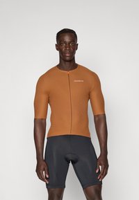 Brun, ribbet, kortermet sykkeltrøye med glidelås foran og logo. I kombinasjon med svarte sykkelshorts. Glatt tekstur, tettsittende design.