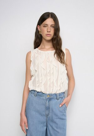 Jeune femme aux longs cheveux bruns portant un chemisier blanc sans manches à volants et un jean bleu taille haute, debout avec une main dans la poche.