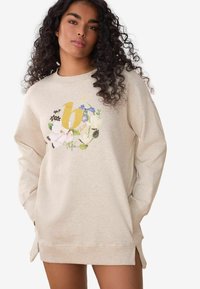 Beżowy sweatshirt z kwiatowym wzorem i złotą literą 'b' z przodu. Posiada długie rękawy, okrągły dekolt oraz boczne rozcięcia u dołu.