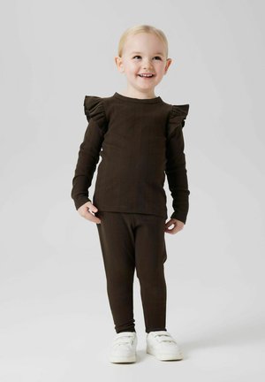 Ein lächelndes kleines Kind, das ein dunkelbraunes Outfit mit Rüschen an den Schultern und weiße Sneakers trägt, steht vor einem hellgrauen Hintergrund.