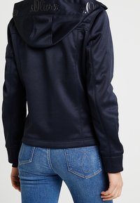 Mörkblå huvjacka i slätt tyg, med präglat varumärke på kragen och synliga sömnadsdetaljer. Matcher med jeans i denim.