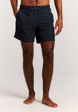 Homme debout pieds nus portant un short de bain noir avec des poches latérales et un cordon bleu sur un fond clair.