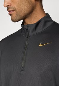 Crna dugosleeve majica Nike s polu-zatvaračem na ovratniku, teksturirana tkanina i zlatni logo na lijevoj strani prsnog koša. Glatka završnica, dizajn koji se prilagođava tijelu.