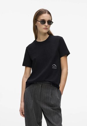 Femme portant des lunettes de soleil noires, un t-shirt noir à manches courtes avec un petit texte blanc, et un pantalon gris à fines rayures, les mains dans les poches.