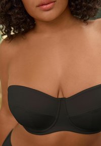 Soutien-gorge bandeau noir en tissu lisse, avec des bonnets structurés et des coutures. Il dispose d'une bande sans couture et d'un design légèrement structuré pour un soutien optimal.