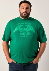 Tyrkysové tričko s krátkým rukávem a grafickým potiskem orla a textem "DENIM BRAND EST. 2007 NEW YORK CITY." Tkanina vypadá hladce, má kulatý výstřih.